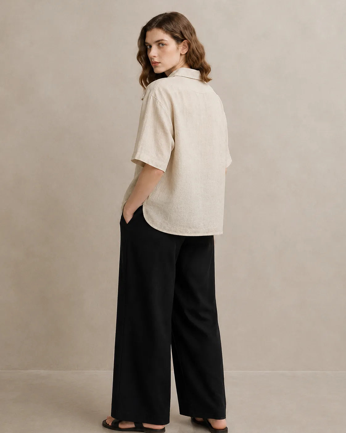Three-quarter back view of a woman wearing an oversized linen beige V-neck shirt.