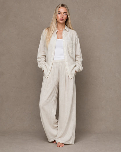 Full-length shot of a woman wearing a beige linen shirt open over trousers in a matching tone.
