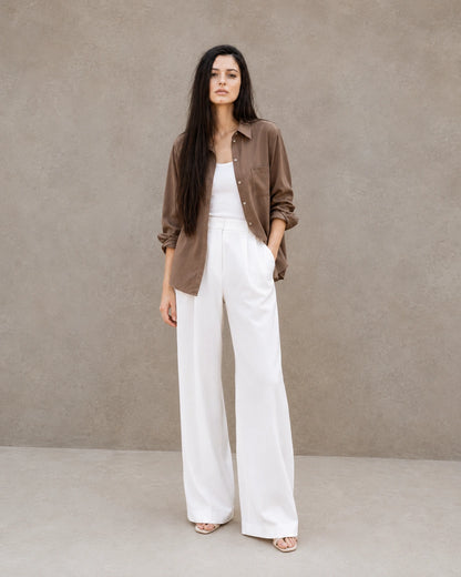 Full-length shot of a woman wearing an open brown lyocell-linen shirt, styled with wide-leg white trousers.