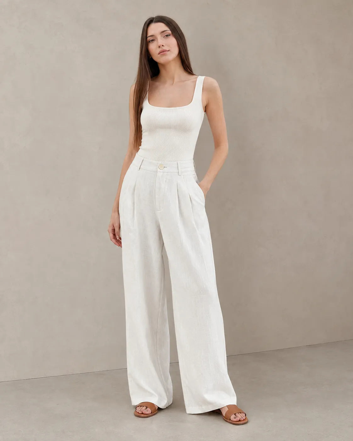 Model wearing white wide-leg summer trousers in a cotton-linen-polyester blend with a pleated high waist, styled with a ribbed white tank top, front view.