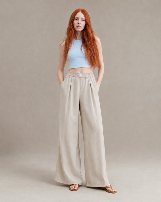 Model draagt een gedrapeerde linnen broek dames in abrikooskleurige linnen-polyestermix met elastische, geknoopte taille, gestyled met een lichtblauw croptopje, vooraanzicht.