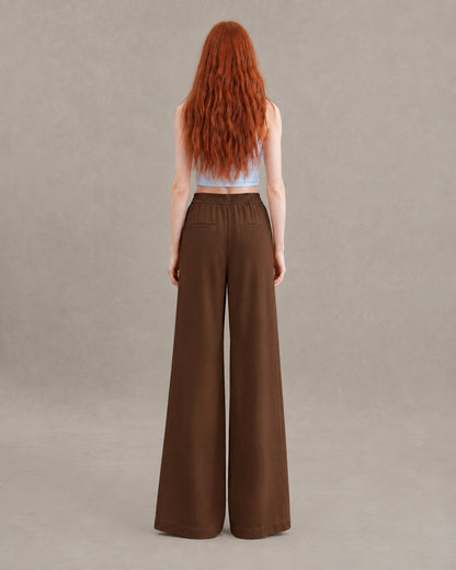 Achteraanzicht van model in een bruine gedrapeerde linnen broek dames in linnen-polyestermix met elastische taille en steekzakken.