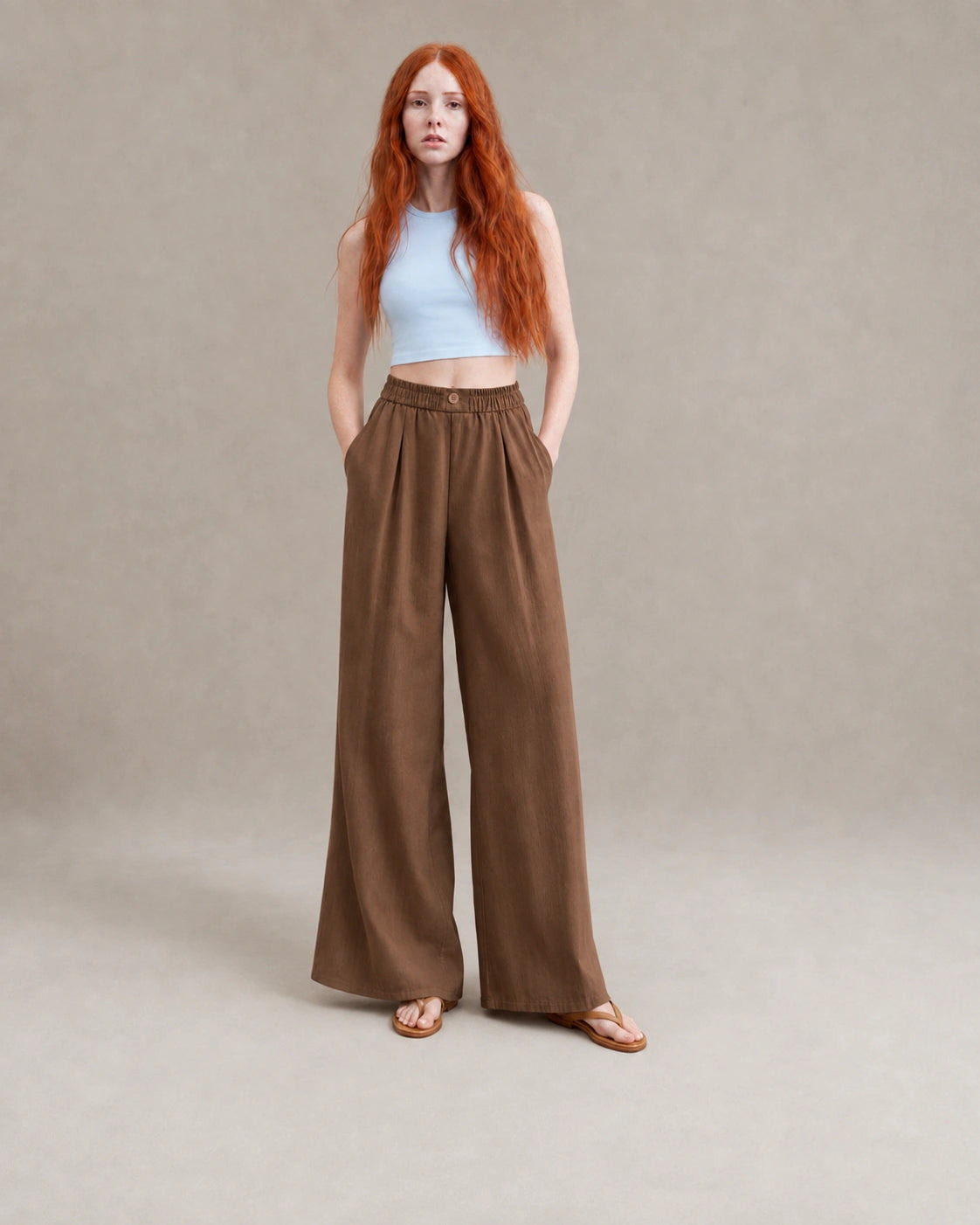 Model draagt een gedrapeerde linnen broek dames in bruine linnen-polyestermix met elastische, geknoopte taille, gestyled met een lichtblauw croptopje, vooraanzicht.