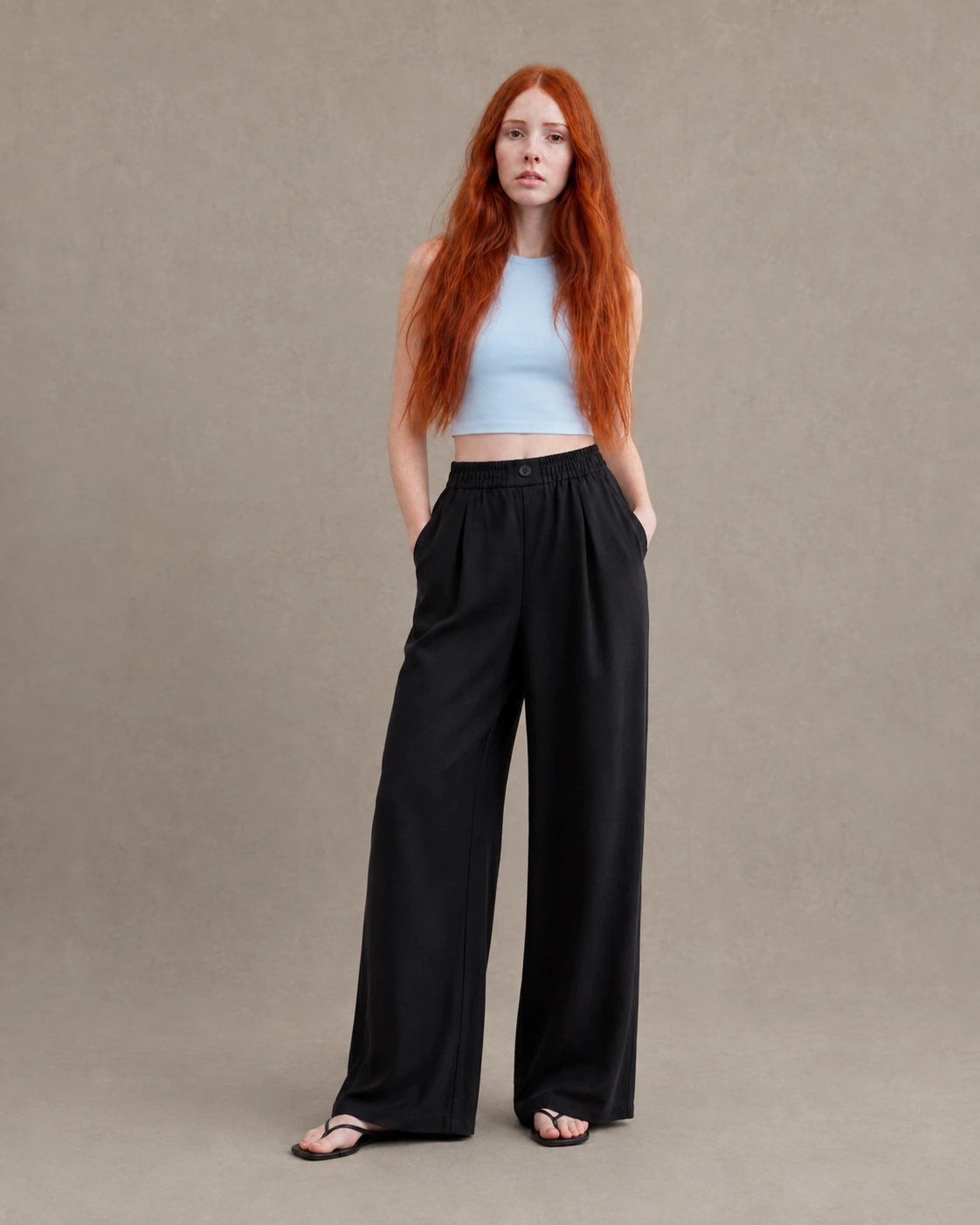 Model draagt een gedrapeerde linnen broek dames in zwarte linnen-polyestermix met elastische, geknoopte taille, gestyled met een lichtblauw croptopje, vooraanzicht.