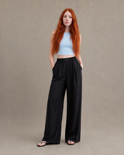 Model draagt een gedrapeerde linnen broek dames in zwarte linnen-polyestermix met elastische, geknoopte taille, gestyled met een lichtblauw croptopje, vooraanzicht.