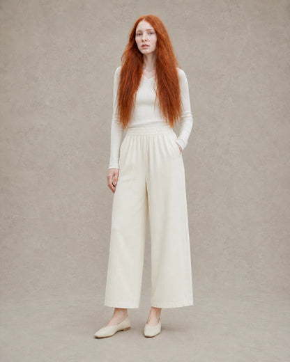 Model draagt een ingekorte wijde broek dames in gebroken wit tencel-linnenmix met elastische taille, gestyled met een crème geribbelde top, vooraanzicht.