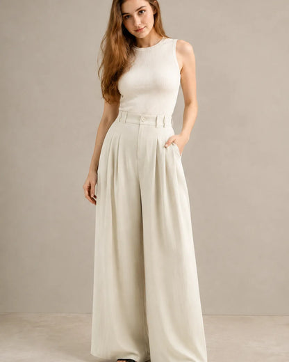 Model wearing white wide-leg trousers in a tencel-linen blend with a draped finish and pleated high waist, styled with an ivory ribbed tank top, three-quarter front view.