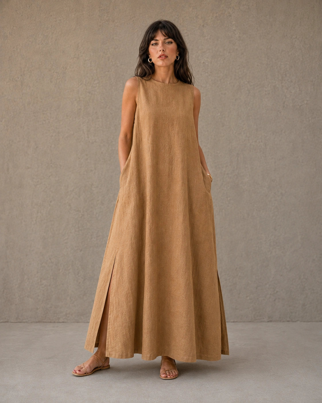 Vrouw draagt een mouwloze linnen A-lijn jurk in camel, met zicht op de splits in de zijzoom, vooraanzicht.