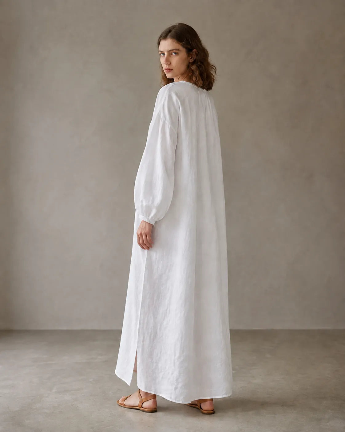 Back view of a white cotton-linen dress with long sleeves and a side hem slit, shown on a model.